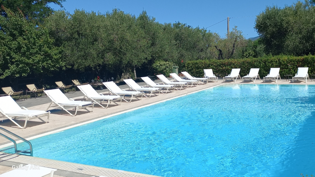 Piscina all'aperto con lettini bianchi, circondata da alberi e cielo sereno.