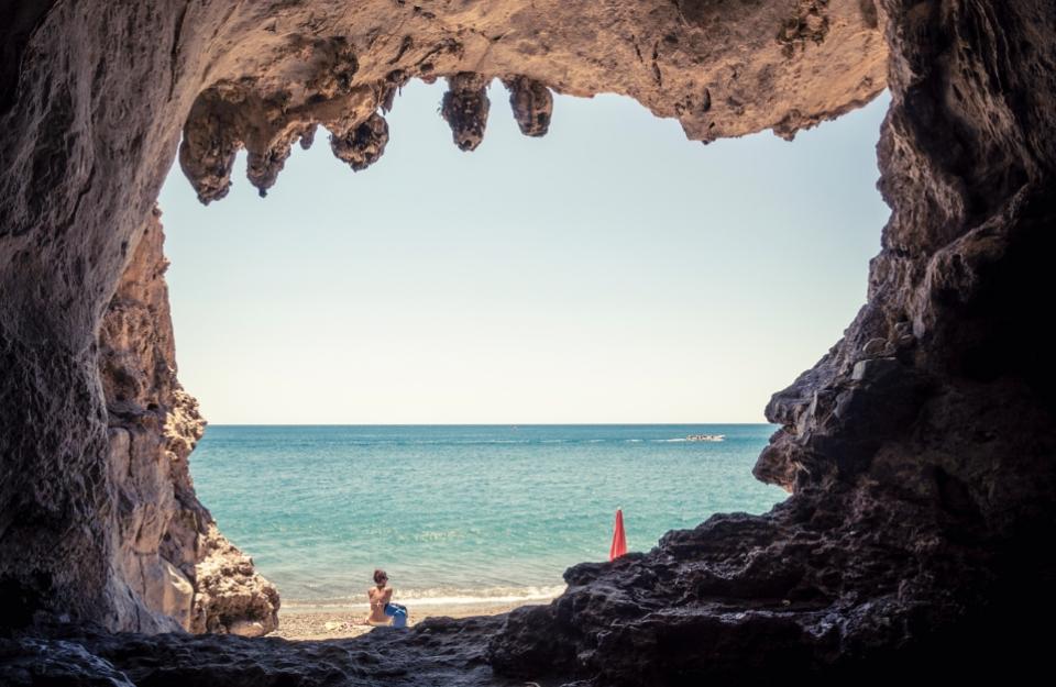 Vista da una grotta sulla spiaggia, con una persona seduta vicino al mare.