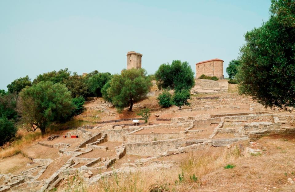 Rovine antiche con torre e edificio in pietra tra alberi e vegetazione.