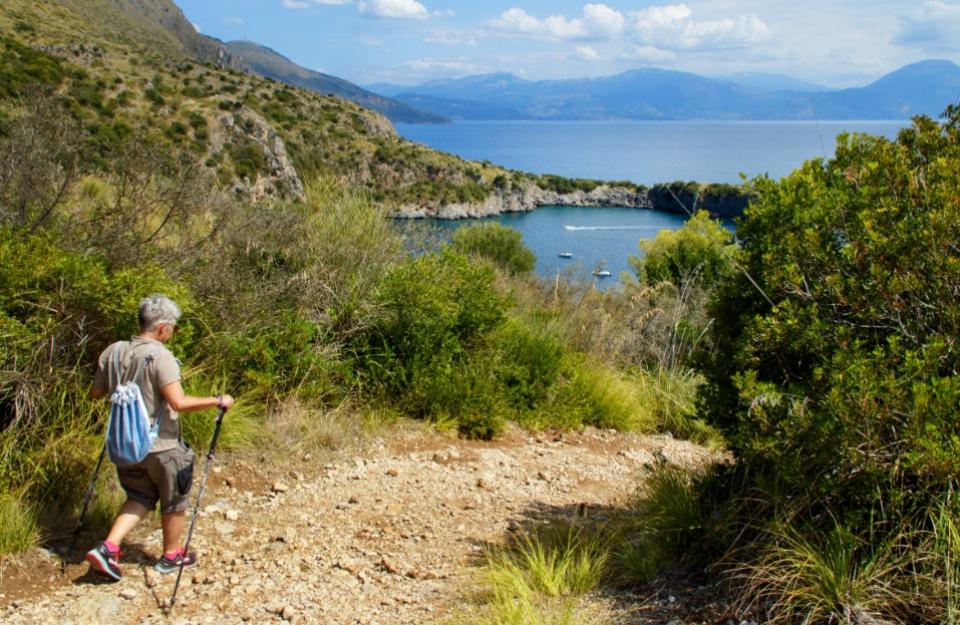 Escursionista su sentiero costiero con vista mare e montagne sullo sfondo.